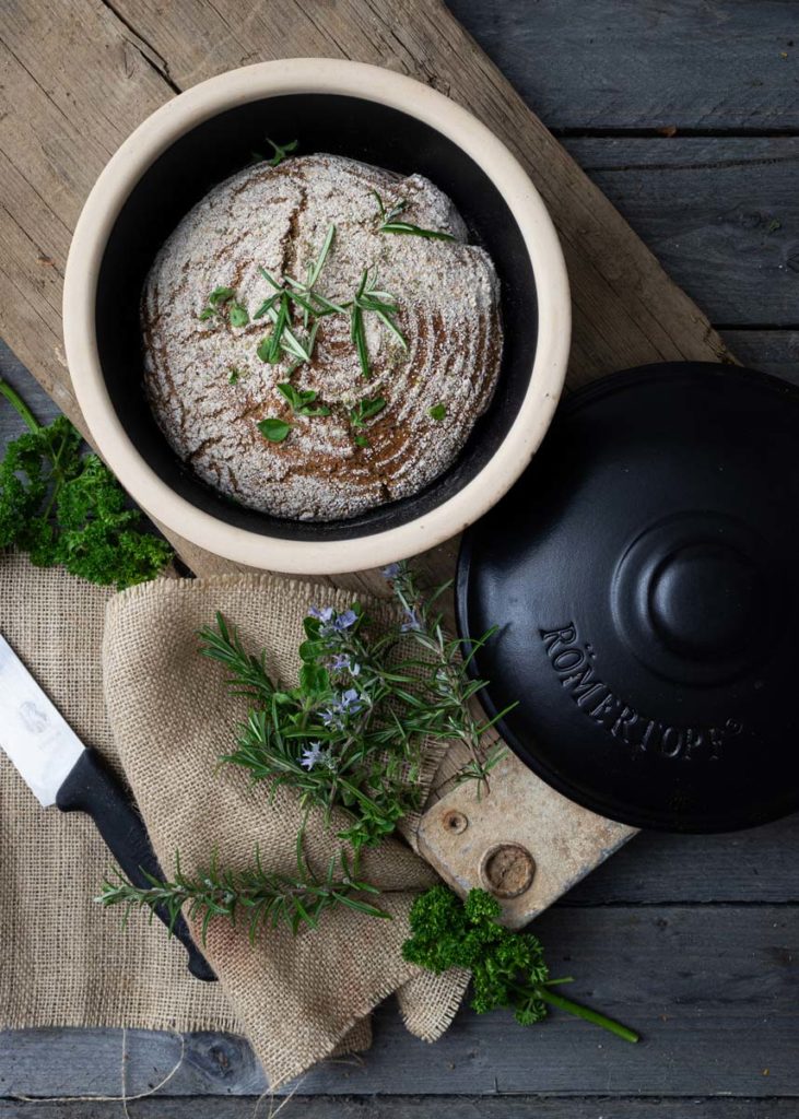 Rundes Brot in einem Römertopf, dekoriert mit frischen Kräutern, auf einem Holzbrett und Jutesack neben einem Messer.