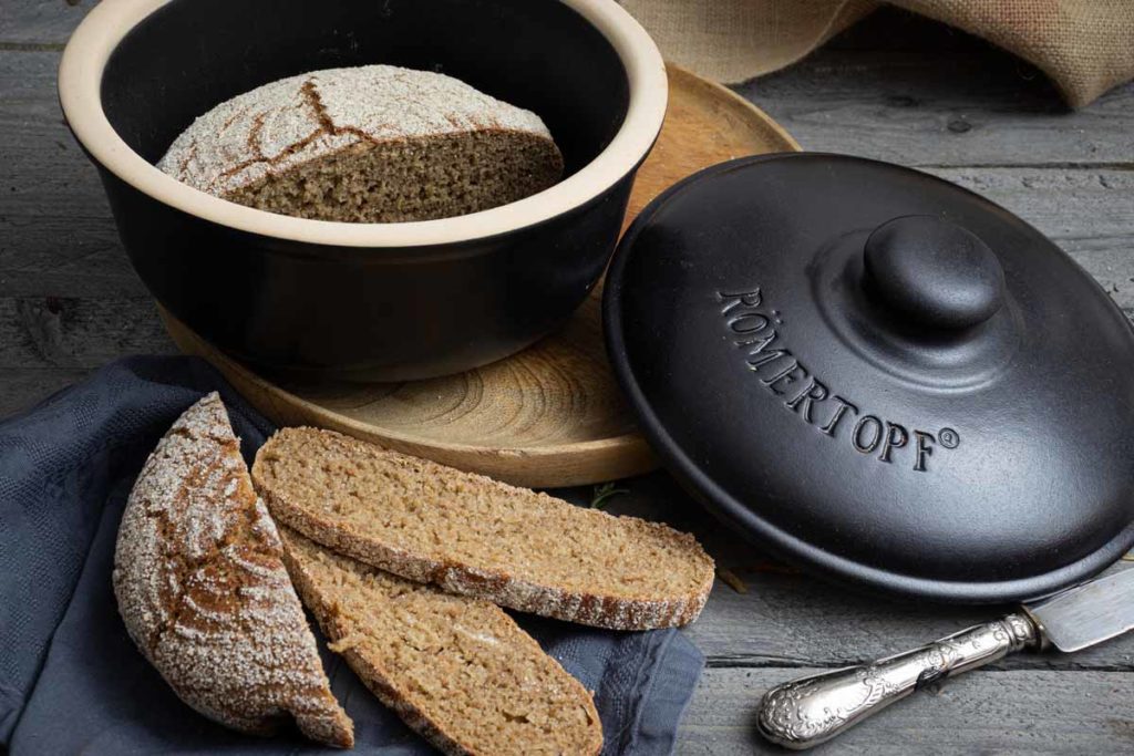 Brot im Römertopf, aufgeschnitten auf Holzbrett, daneben schwarzer Deckel und Messer auf grauem Tuch.