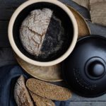 Brotlaib im Topf mit zwei Brotscheiben daneben, auf Holztisch mit dunklem Tuch und Leinenstoff.