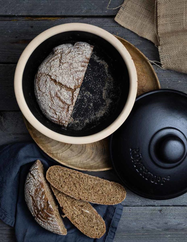Brotlaib im Topf mit zwei Brotscheiben daneben, auf Holztisch mit dunklem Tuch und Leinenstoff.