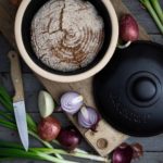 Rundes Brot in einem Topf auf Holzbrett, umgeben von Zwiebeln, Frühlingszwiebeln und einem Messer.