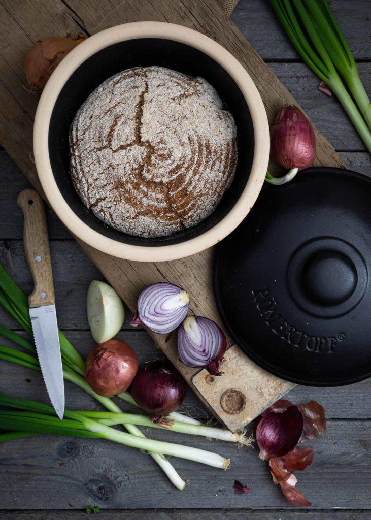 Rundes Brot in einem Topf auf Holzbrett, umgeben von Zwiebeln, Frühlingszwiebeln und einem Messer.