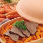 Braten mit Karotten und Petersilie in einem Römertopf, umgeben von frischen Karotten auf einem Holztisch.
