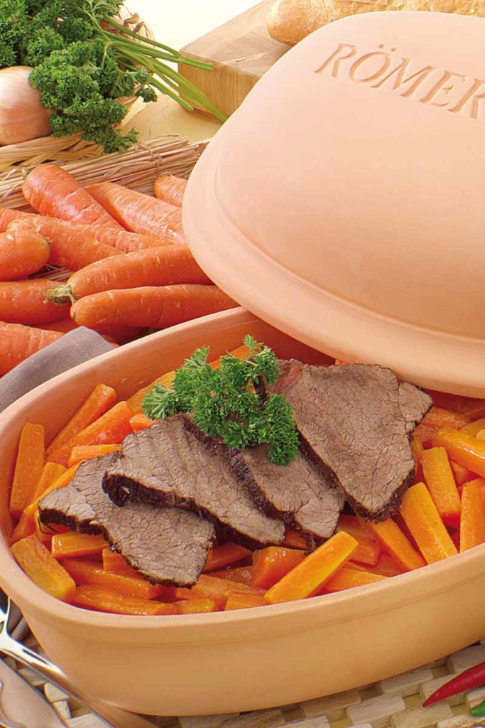 Braten mit Karotten und Petersilie in einem Römertopf, umgeben von frischen Karotten auf einem Holztisch.