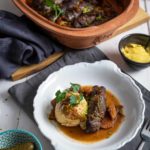 Weißer Teller mit Fleischroulade, Serviettenknödel, Soße und Petersilie. Im Hintergrund Gemüse im Römertopf, Senf und Gurken.