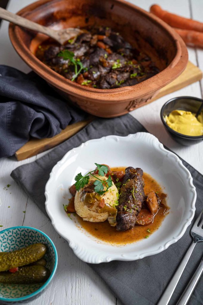 Weißer Teller mit Fleischroulade, Serviettenknödel, Soße und Petersilie. Im Hintergrund Gemüse im Römertopf, Senf und Gurken.