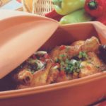 Bratenschenkel im Tontopf mit Paprika und Frühlingszwiebeln, dekoriert mit frischen Kräutern. Brot und Paprika im Hintergrund.