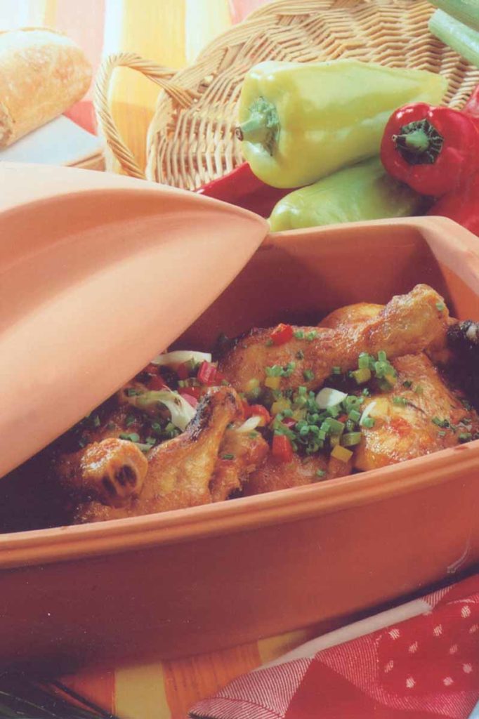 Bratenschenkel im Tontopf mit Paprika und Frühlingszwiebeln, dekoriert mit frischen Kräutern. Brot und Paprika im Hintergrund.