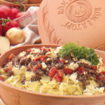 Nudelauflauf mit Hackfleisch, Tomaten und Käse in einem Römertopf. Im Hintergrund Brot, Zwiebeln und Tomaten.