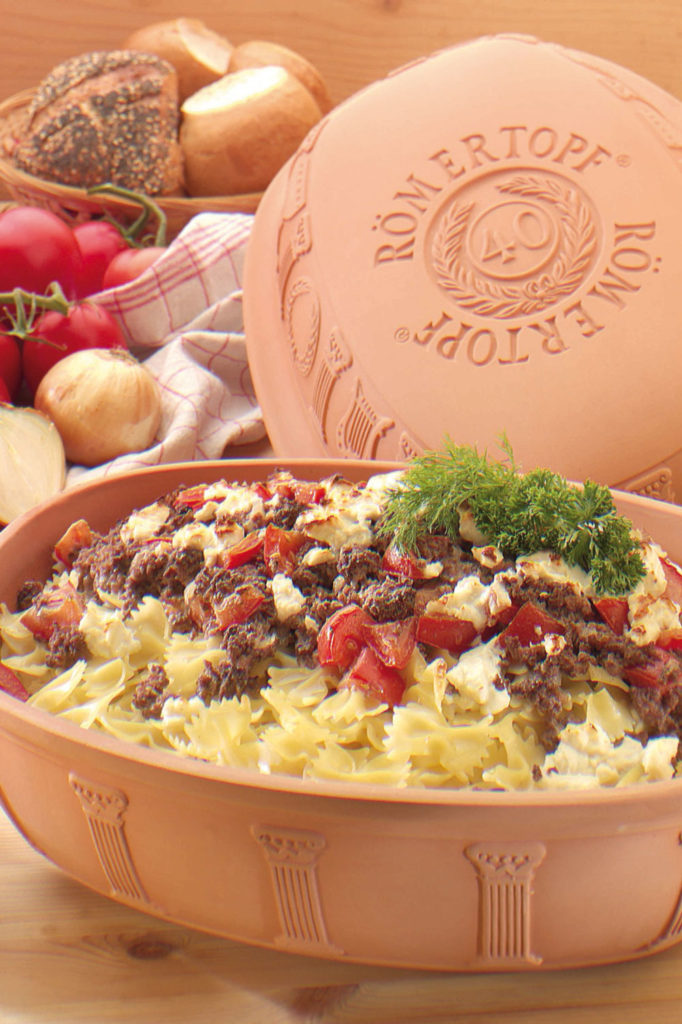 Nudelauflauf mit Hackfleisch, Tomaten und Käse in einem Römertopf. Im Hintergrund Brot, Zwiebeln und Tomaten.