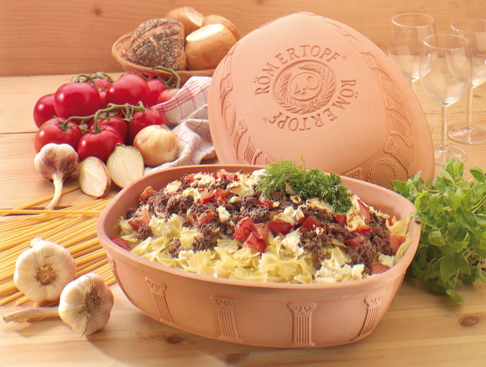 Keramiktopf mit Auflauf aus Nudeln, Hackfleisch, Tomaten und Käse. Umgeben von Brot, Kräutern und Gemüse.