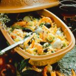 Reistopf mit Meeresfrüchten in einem traditionellen Tontopf serviert, umgeben von Gewürzen und einem Glas Rotwein.