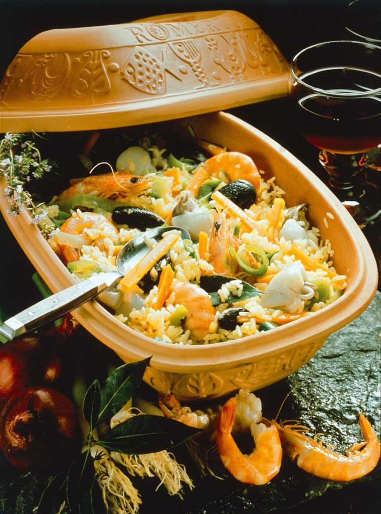 Reistopf mit Meeresfrüchten in einem traditionellen Tontopf serviert, umgeben von Gewürzen und einem Glas Rotwein.