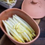 Weißer Spargel in einer Tonform mit Zitronenscheiben und Pfeffer, neben einem Korb mit frischem Spargel.