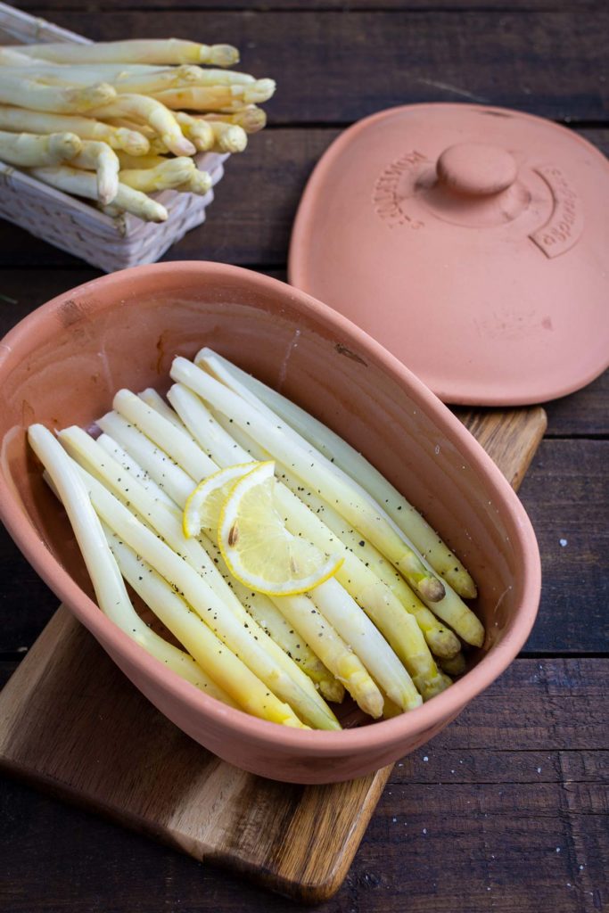 Weißer Spargel in einer Tonform mit Zitronenscheiben und Pfeffer, neben einem Korb mit frischem Spargel.