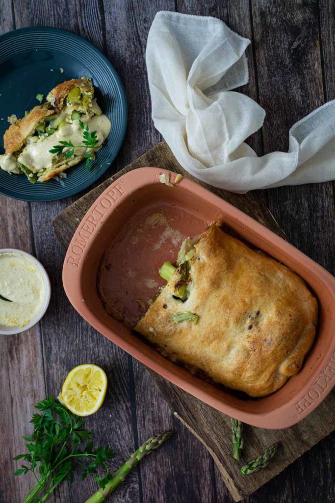 Terrakotta-Backform mit gebackenem Gericht. Ein Stück liegt auf einem Teller. Petersilie und Spargel als Garnitur.