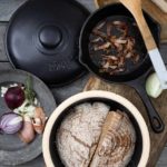 Brot im Tontopf, Speck in Pfanne, Zwiebeln und Schinken auf Teller, Holzlöffel und frische Kräuter auf Holztisch.