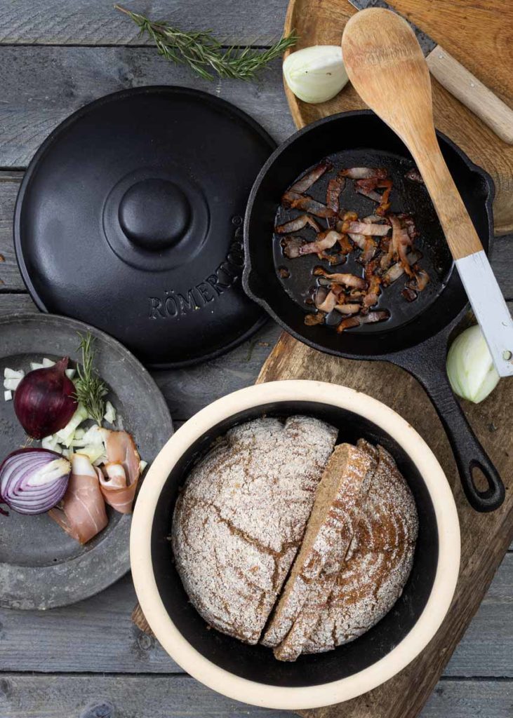Brot im Tontopf, Speck in Pfanne, Zwiebeln und Schinken auf Teller, Holzlöffel und frische Kräuter auf Holztisch.