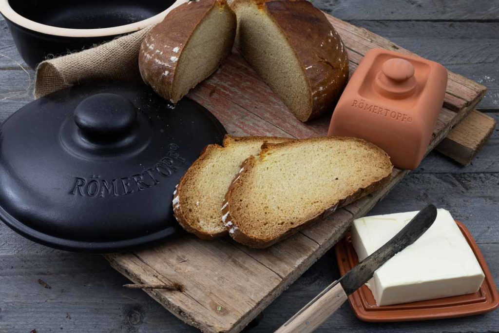 Roggenbrot, Tonschale von Römertopf, Butter und Messer auf Holzbrett. Rustikale Essenszene.