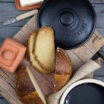 Geschnittener Laib Brot und Butter auf Holztisch mit einem schwarzen Römertopf und zwei Tongefäßen.
