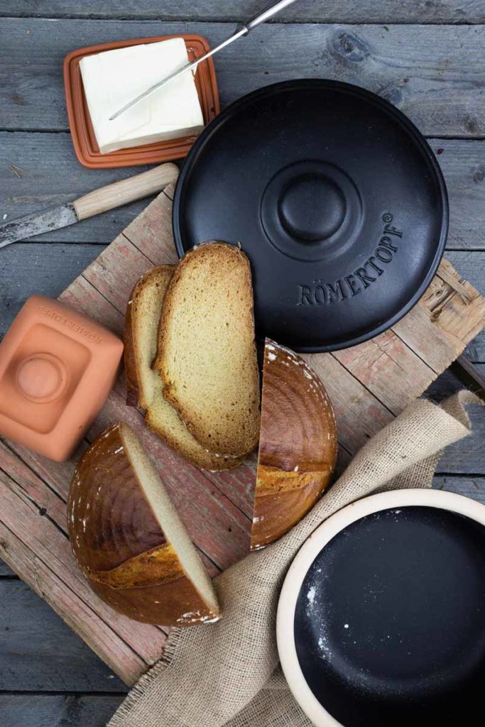 Geschnittener Laib Brot und Butter auf Holztisch mit einem schwarzen Römertopf und zwei Tongefäßen.