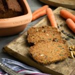 Karottenbrot, geschnitten auf Holzbrett mit Möhren und Nüssen, im Hintergrund Römertopf und silberner Löffel.