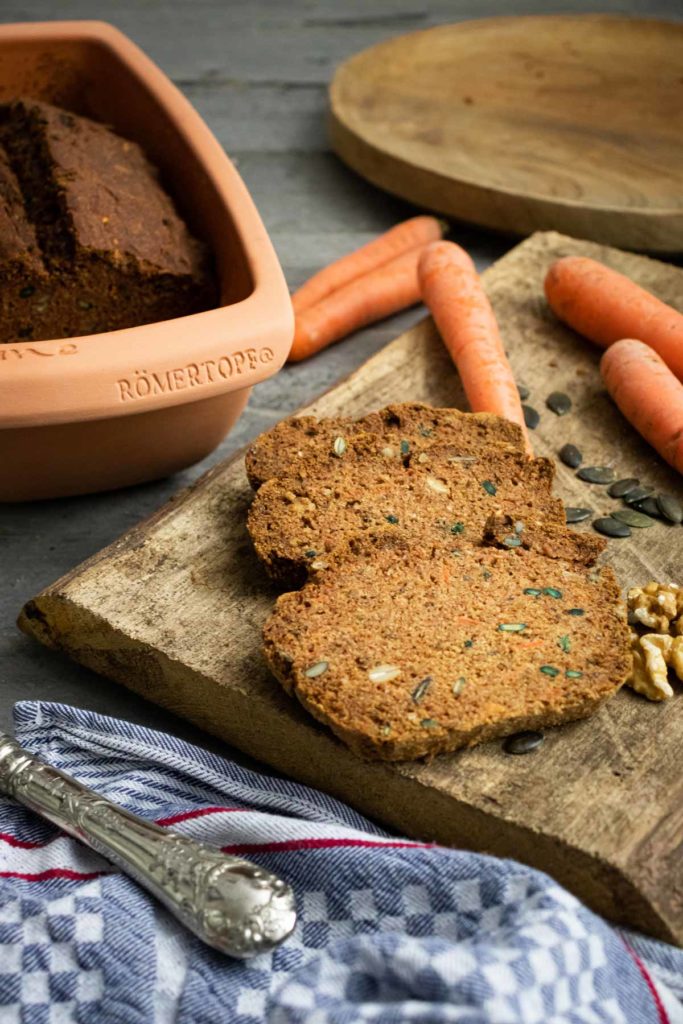 Karottenbrot, geschnitten auf Holzbrett mit Möhren und Nüssen, im Hintergrund Römertopf und silberner Löffel.