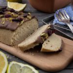 Kuchen mit Schokoglasur, Pistazien und Beeren auf Holzbrett, daneben Zitronenscheiben und ein Tontopf.