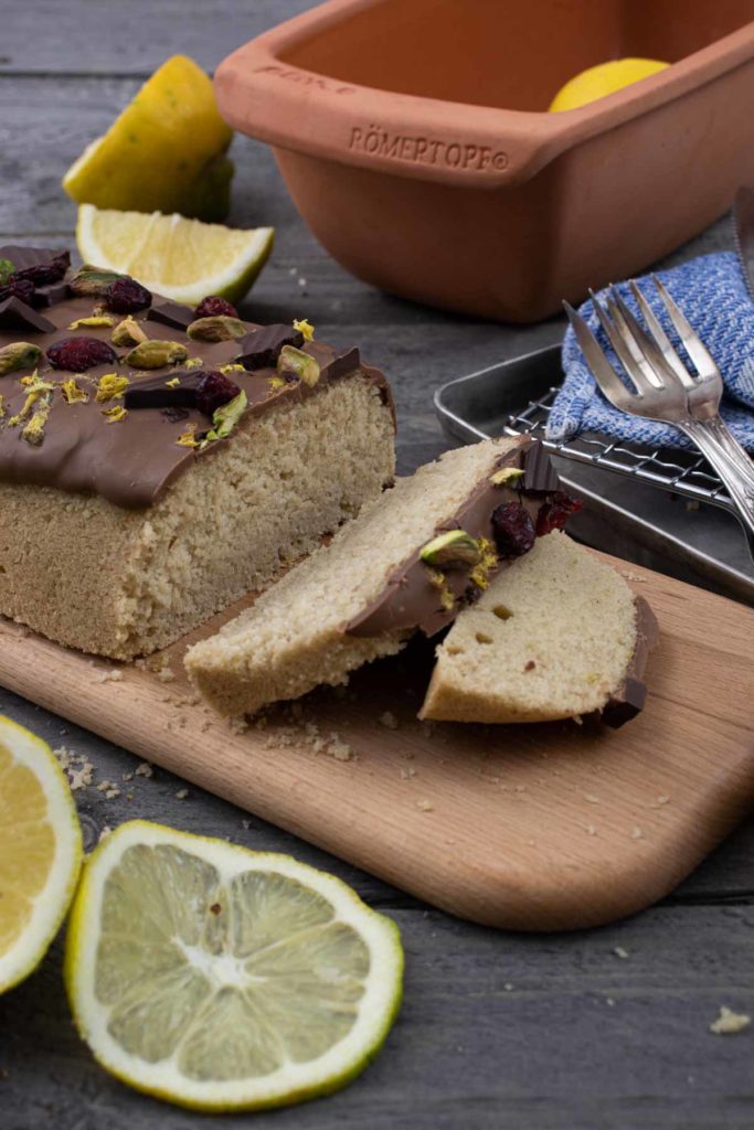 Kuchen mit Schokoglasur, Pistazien und Beeren auf Holzbrett, daneben Zitronenscheiben und ein Tontopf.