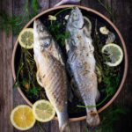 Zwei gebratene Fische in einer Pfanne mit Zitronenscheiben, Knoblauch, Kräutern und Gewürzen auf Holzuntergrund.