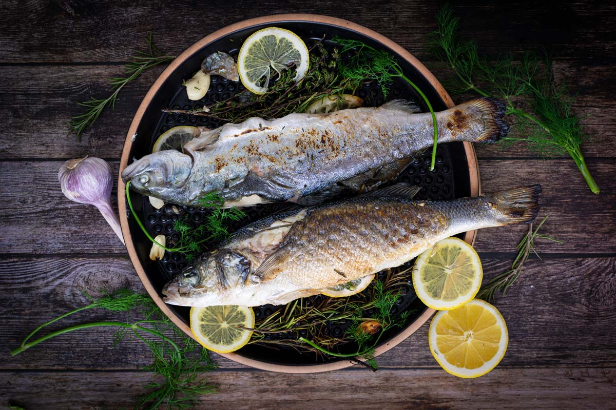 Zwei gegrillte Fische auf Kräutern und Zitronenscheiben, dekoriert mit Knoblauch und Dill auf einem Holztisch.