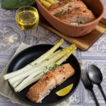 Gegrillter Lachs mit weißem Spargel auf schwarzem Teller, serviert neben einem Glas Weißwein und Besteck.