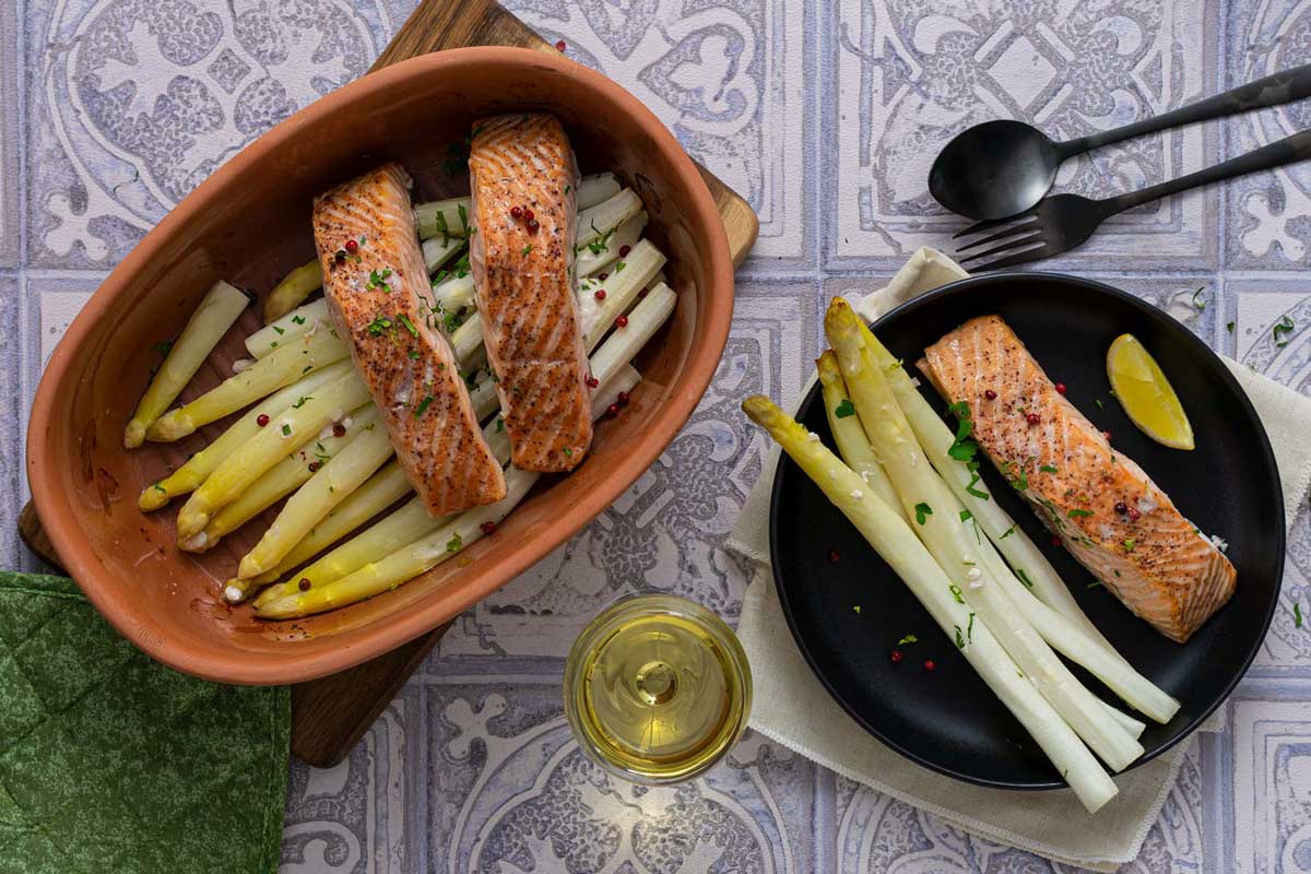 Gebratener Lachs und Spargel auf einem schwarzen Teller, daneben Lachs und Spargel in einer Terrakotta-Schale, mit Zitrone.
