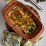 Auflauf mit Spargel und Käse in brauner Keramikform auf Holzbrett, daneben Zitrone, Spargel, Spatel, Petersilie und Tuch.