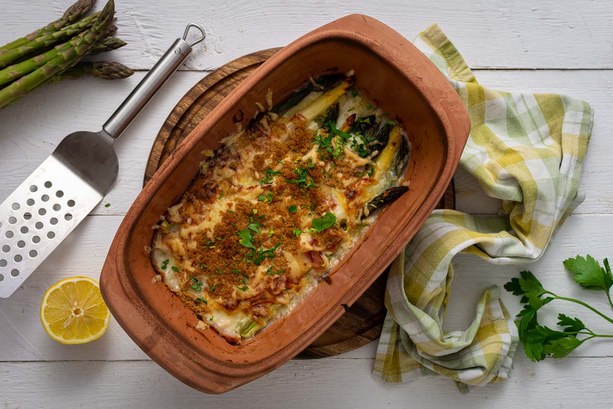 Überbackener Spargel in einer Terrakotta-Form neben Zitronenhälfte und Spatel auf weißem Tisch.