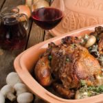 Gebackenes Hähnchen im Römertopf mit Champignons, Rotwein und Zwiebeln auf einem Holztisch.