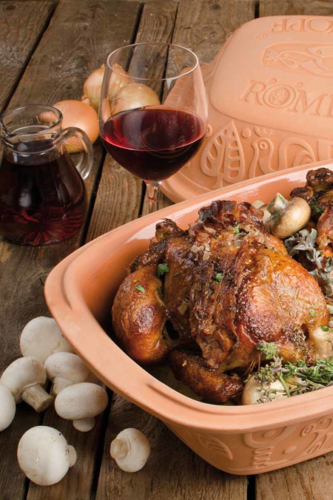 Gebackenes Hähnchen im Römertopf mit Champignons, Rotwein und Zwiebeln auf einem Holztisch.