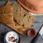 Rustikale Brotscheiben auf Holzbrett, daneben Joghurt, Marmelade und ein Messer, Romertopf im Hintergrund.