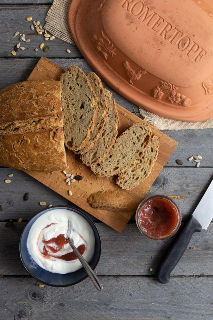 Rustikale Brotscheiben auf Holzbrett, daneben Joghurt, Marmelade und ein Messer, Romertopf im Hintergrund.