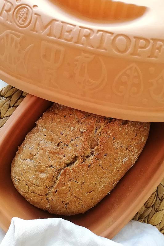 Brotlaib in einem teilweise geöffneten Römertopf aus Ton auf einem geflochtenen Untergrund.
