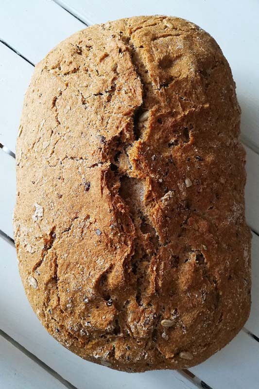 Vollkornbrotlaib mit knuspriger Kruste und dunklen Saaten auf einem weißen Holztisch.