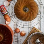 Gugelhupf auf einem Kuchengitter, mit Eiern, Backpulver und geschmolzener Schokolade auf einem Holztisch arrangiert.