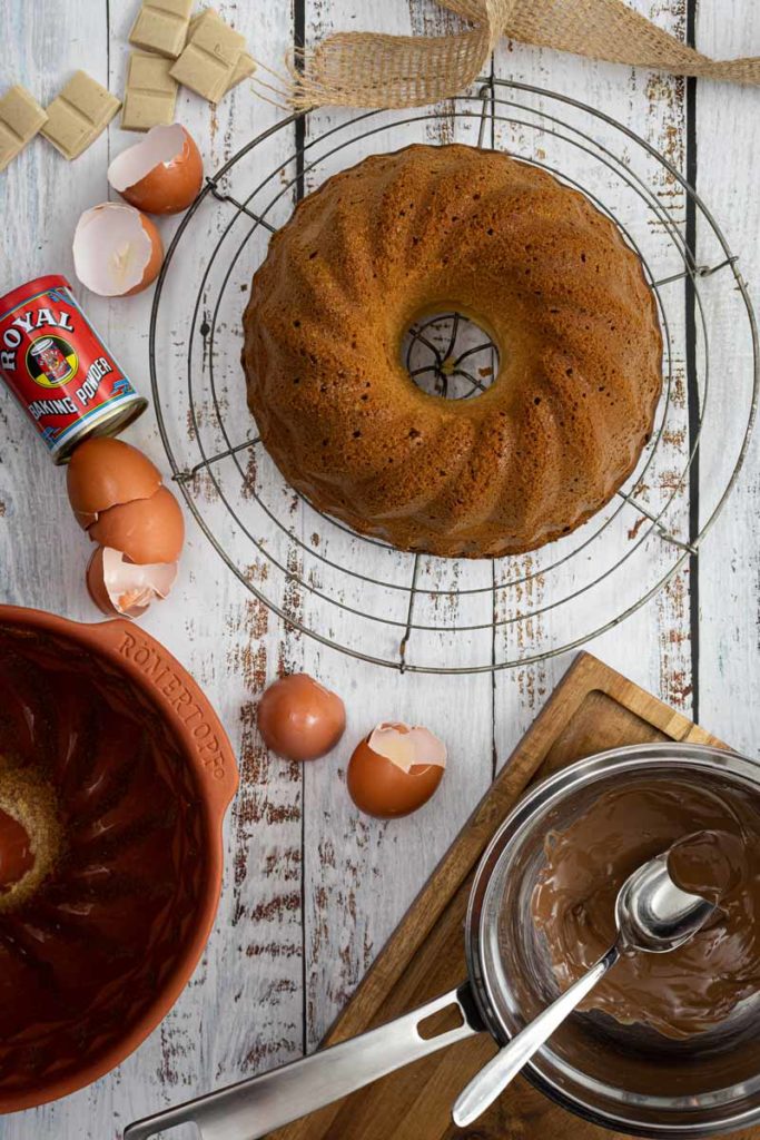Gugelhupf auf einem Kuchengitter, mit Eiern, Backpulver und geschmolzener Schokolade auf einem Holztisch arrangiert.