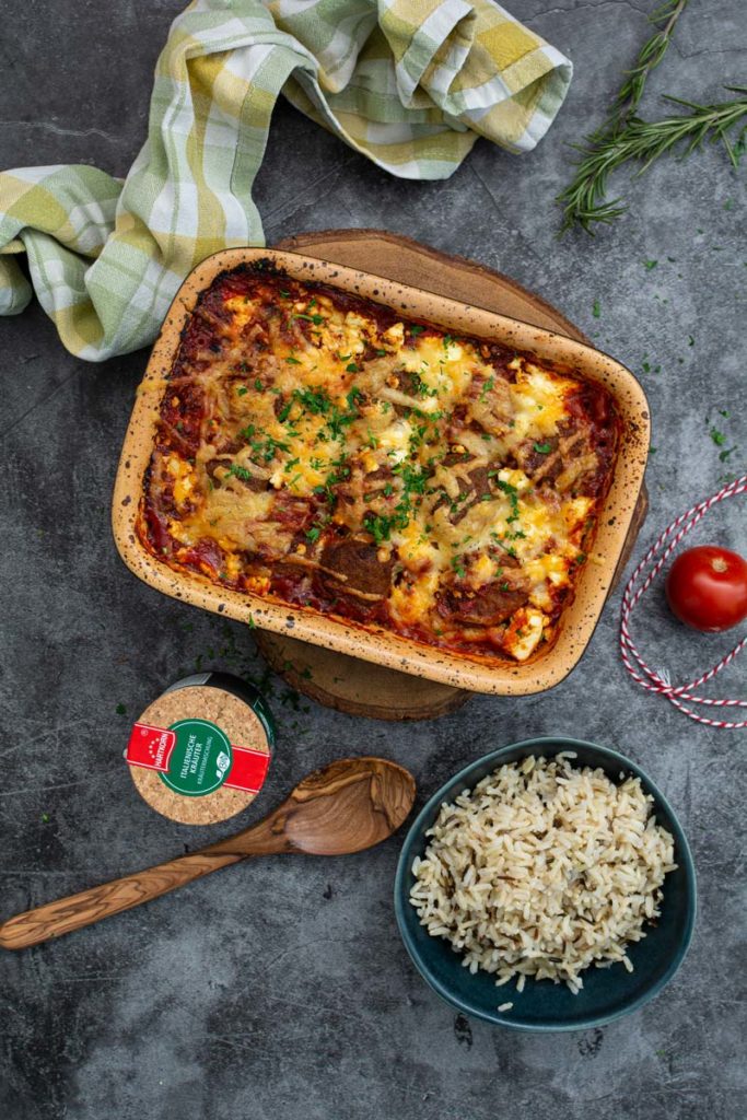 Auflaufform mit überbackenem Gericht, daneben Schüssel mit Reis, Holzlöffel, Tomate, Gewürzdose und Stoffserviette.