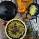 Kartoffelauflauf in einem Topf, umgeben von Kartoffeln, Zwiebeln, Käse und Kräutern, auf einer Holzoberfläche.