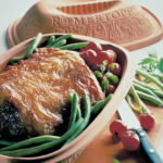Bräter mit Braten, grünen Bohnen und Kirschtomaten. Romertopf-Deckel im Hintergrund auf einem Holzbrett.