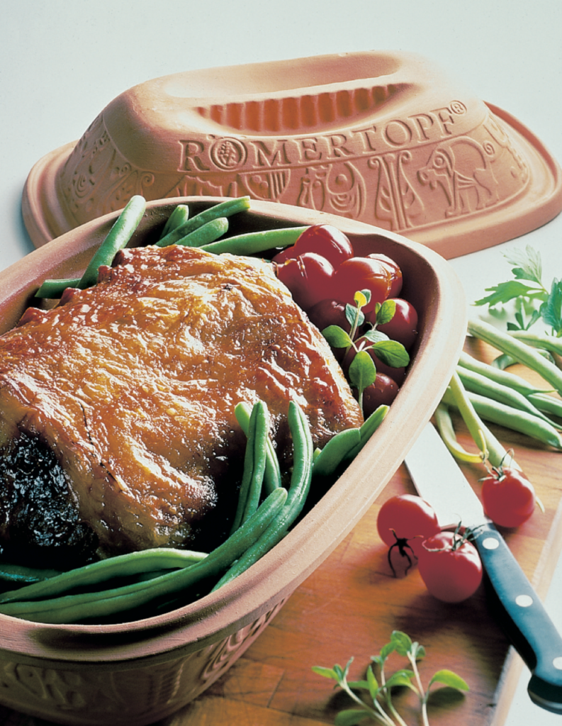 Bräter mit Braten, grünen Bohnen und Kirschtomaten. Romertopf-Deckel im Hintergrund auf einem Holzbrett.