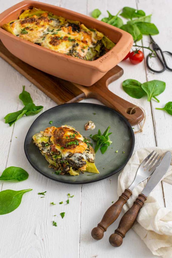 Vegetarische Lasagne auf einem Teller vor einer Auflaufform mit goldbrauner Kruste, umgeben von frischem Spinat.