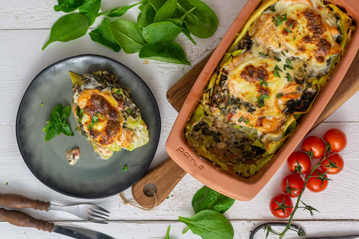 Lasagne mit Spinat und Käse in einer Tonform neben einem Teller, umgeben von Tomaten und frischem Spinat.