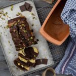 Schokoladen-Pistazien-Kuchen auf einem Blech mit Schokostücken, geschnitten, neben leere Backform und Tuch.